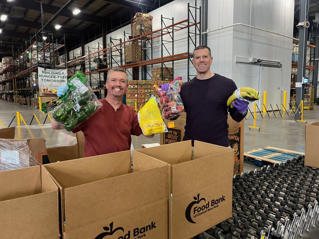 Pat and AJ from HighPoint Advisors, LLC smiling on the volunteer line, holding up fresh produce as they pack boxes for the Food Bank of Central New York.