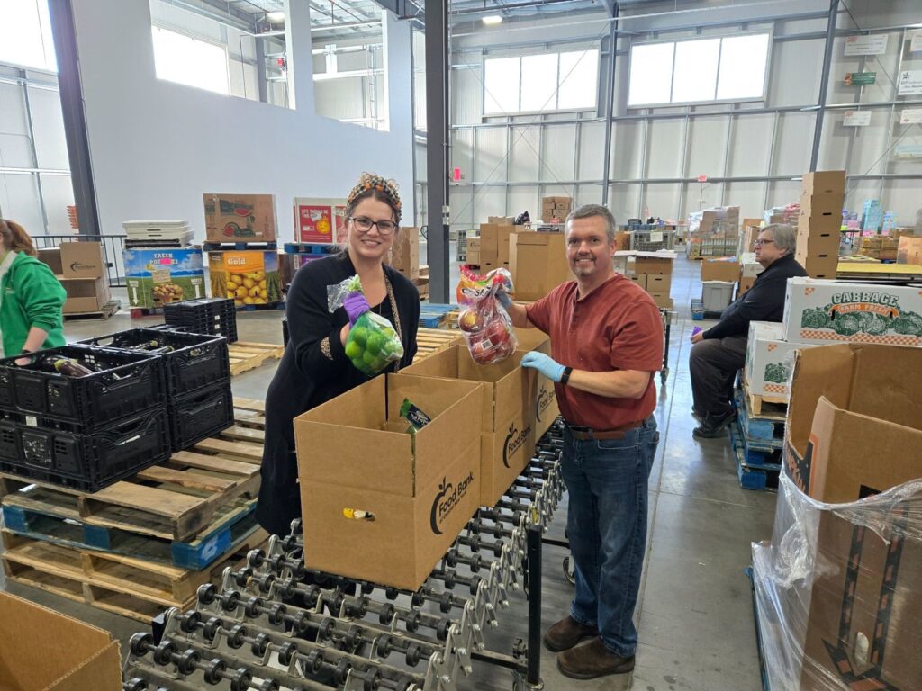 Taylor and Pat from HighPoint Advisors, LLC smiling on the volunteer line, holding up fresh produce as they pack boxes for the Food Bank of Central New York.