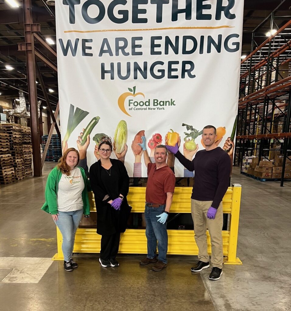 HighPoint Advisors, LLC team posing in front of the Food Bank of Central New York poster at their facilities after volunteering, celebrating their contribution to the community.